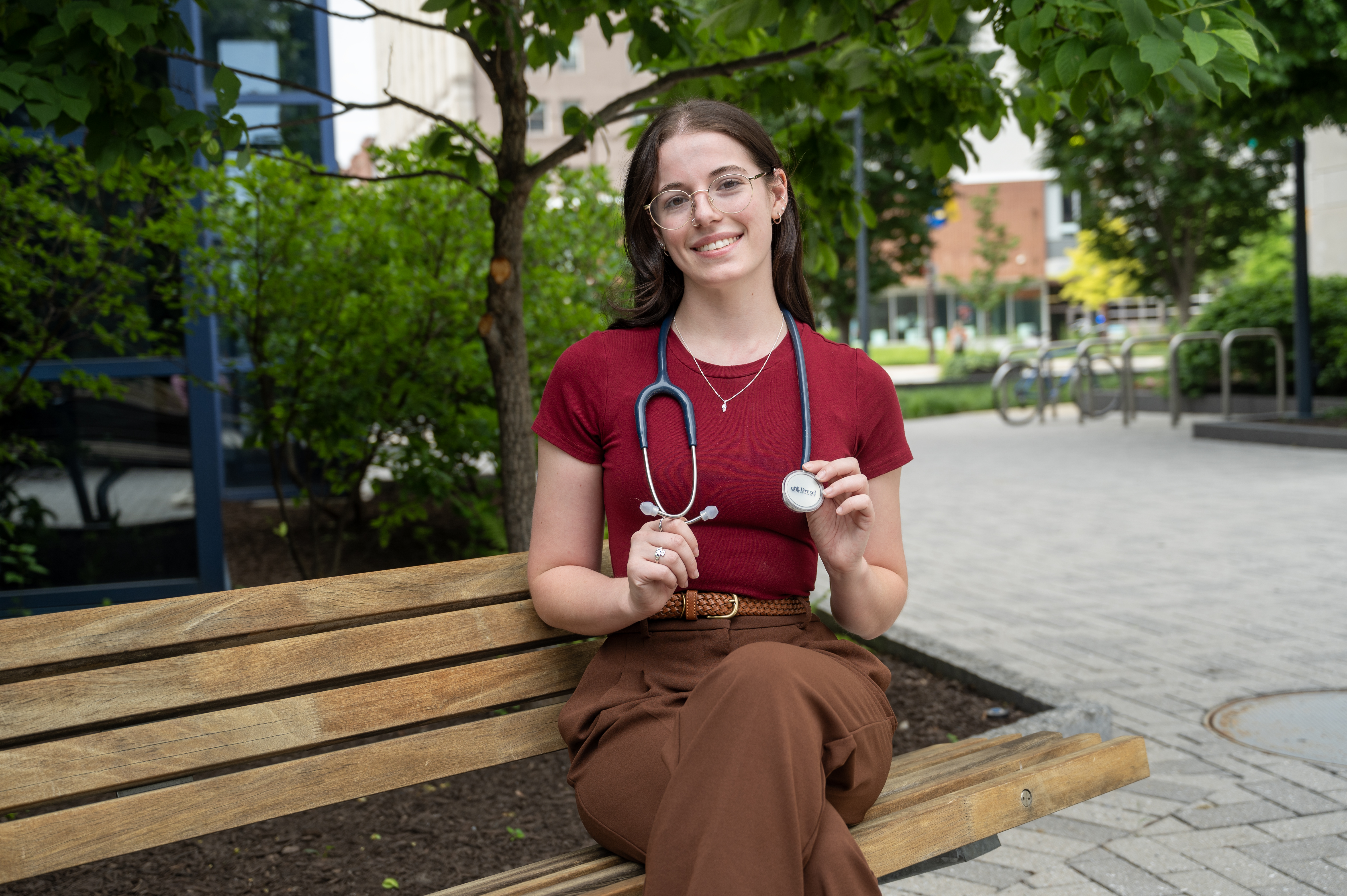 Stephanie Bieder wearing red shirt and brown pants holding stethoscope sitting on bench